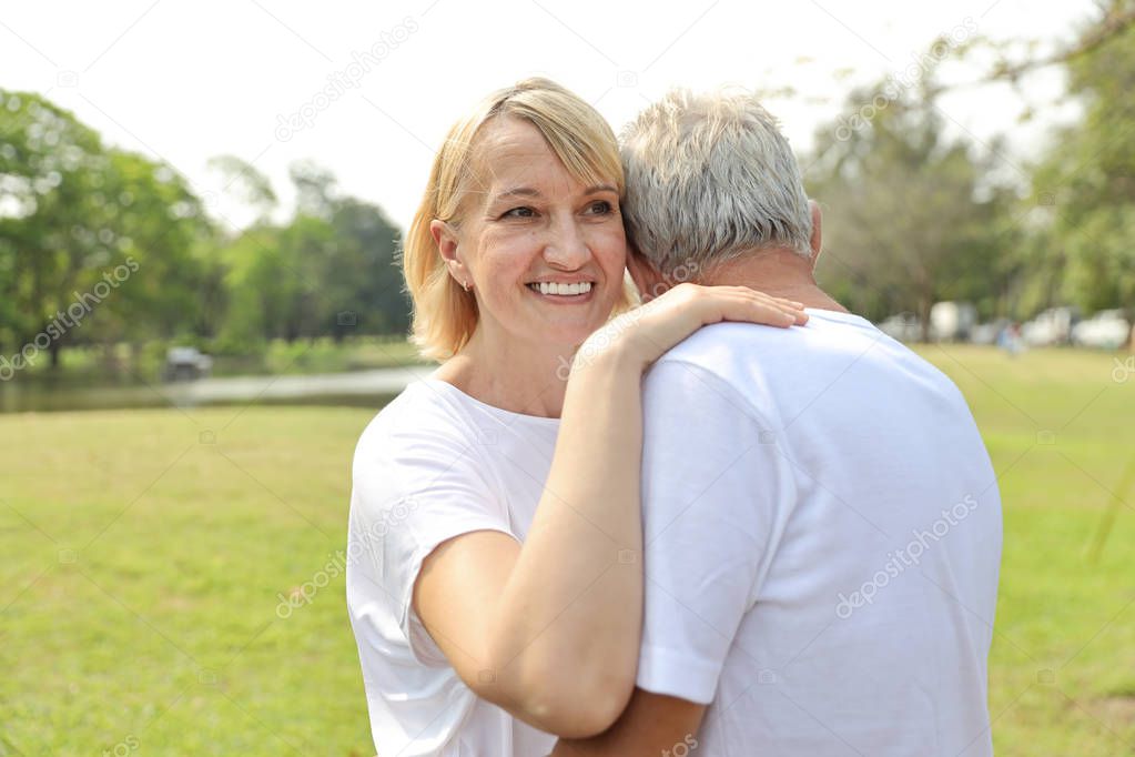 smiling elderly caucasian couple with white shirt and blue jean embracing in park in summer time