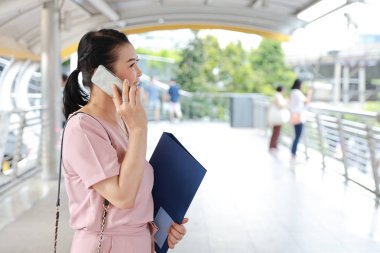 yan görünümü şehir merkezinde yürüyüş yolu ayakta pembe rahat elbise başarılı yaşlı asya lı iş kadını, o cep telefonu kullanarak ve şehir arka plan ile klasör üterek