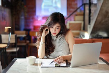 Güzel ve çekici Asyalı kadın, kafeteryada cep telefonu ve dizüstü bilgisayar kullanırken evrak işleriyle uğraşan gri günlük elbiseli uzun saçlı.