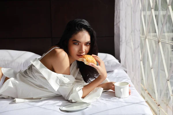 Çekici genç bir kadın sabah kahvaltıda kahve ve kruvasan yiyor. — Stok fotoğraf