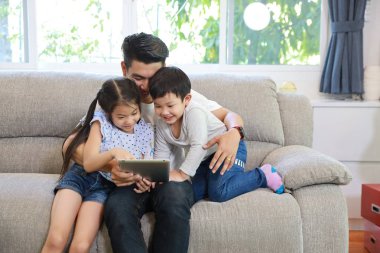 Mutlu Asyalı baba, kızına oturma odasındaki gri koltukta gülen ve gülen yüzlü tablet kullanmayı öğretiyor.)