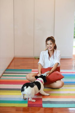 Beyaz gömlek giyen, elinde kırmızı yastıkla oturan ve oturma odasında gülen yüzlü sevimli köpeğiyle oynayan uzun boylu, güzel Asyalı bir kadın.