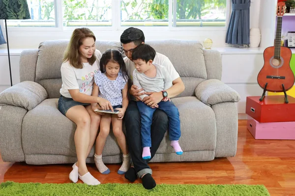 Szczęśliwa azjatycka rodzina uczy swoje dzieci syna i córkę jak używać tabletu siedząc na szarej kanapie w salonie z uśmiechniętymi twarzami — Zdjęcie stockowe