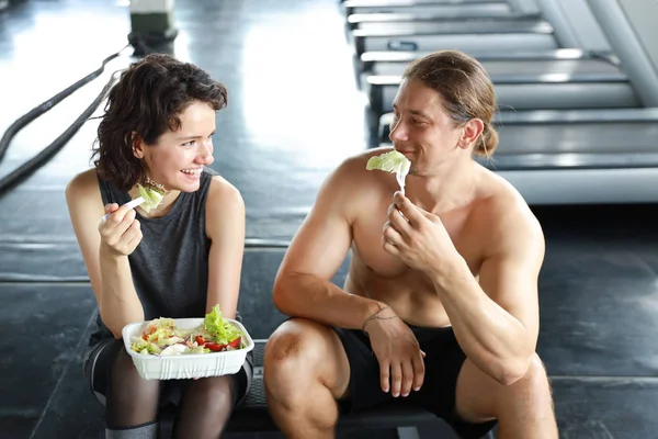 Krásný kavkazský pár fitness muž a žena si odpočinout a jíst salát v tělocvičně po dokončení fitness školení při smíchu a úsměvu — Stock fotografie