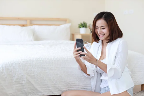 Vista laterale di adulto freelance asiatico donna in bianco camicia lavoro su computer e cellulare in camera da letto con felice sorriso faccia — Foto Stock