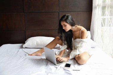 Beyaz tişörtlü yetişkin Asyalı kadın yatak odasında bilgisayar ve cep telefonuyla çalışıyor.