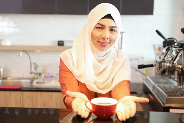 Jovem muslim barista menina fazendo café e mostrando uma xícara de café no balcão com cara sorridente — Fotografia de Stock