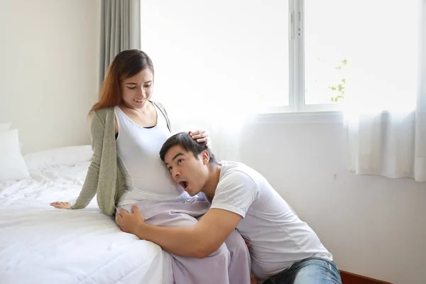 Beau caucasien mari écoute son asiatique enceinte ventre femme avec heureux sourire visage et dire wow sur lit dans chambre — Photo