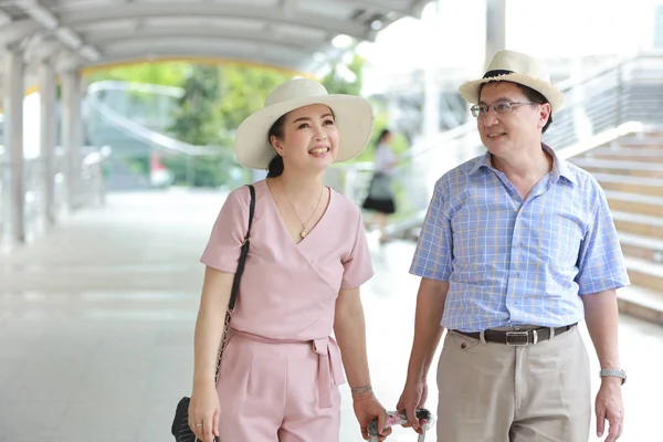 Asiatisches Reisepaar Mann und Frau in rosa und blauem Hemd mit weißem Hut und Gepäck spaziert im Rentenalter glücklich lächelnd durch die Innenstadt — Stockfoto