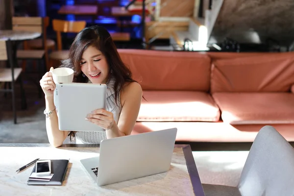 Smuk og attraktiv asiatisk kvinde, langt hår i grå afslappet kjole, der arbejder på bordet og drikker kaffe om morgenen, mens du bruger computer i kaffebar - Stock-foto