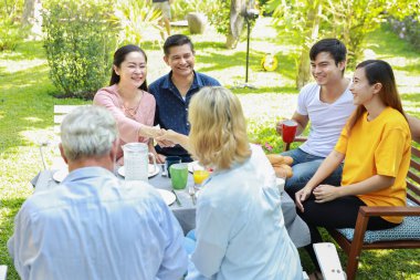 Güneşli bir günde arka bahçedeki kahvaltı masasında oturan mutlu çoklu etnik aile, gülümseyen bir suratla, Asyalı anne ve anne tokalaşırken ve selamlaşırken.