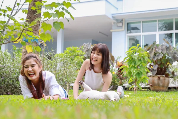 Vacker två unga asiatiska kvinna bär vit skjorta som ligger på gräs och leker med sin söta hund med glada och leende ansikte i trädgården med gröna träd. (vänskapsbegrepp) — Stockfoto