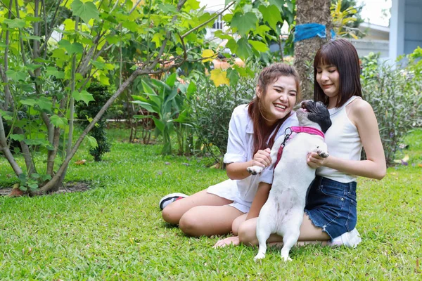 美しい2人の若いアジアの女性が白いシャツを着て草の上に座って、緑の木々と庭で幸せと笑顔で彼女のかわいい犬と遊んでいます。（友情の概念） — ストック写真