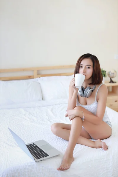 Pleine longueur plan de belle femme asiatique adulte en robe blanche assis sur le lit dans la chambre à coucher tout en utilisant l'ordinateur et boire du café — Photo