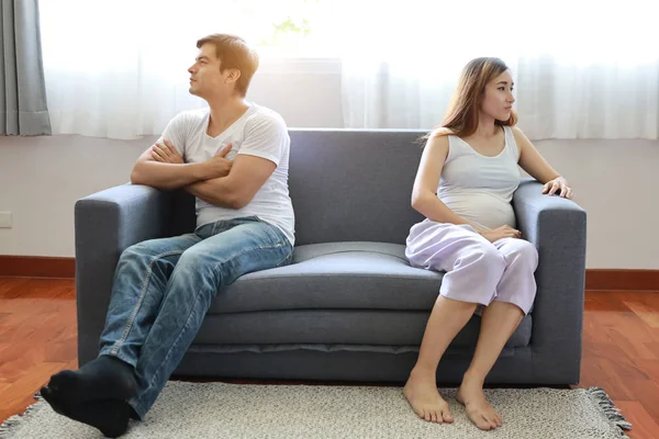 full shot asian pregnant wife and caucasian husband sitting on sofa in living room with angry motion with each other and unhappy face