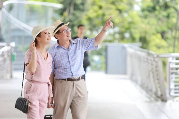 亚洲旅行者夫妇夫妇身穿粉色和蓝色衬衫，头戴白色漂亮的帽子，背着行李，走在市中心，他指着一些带有右复制空间的东西 — 图库照片