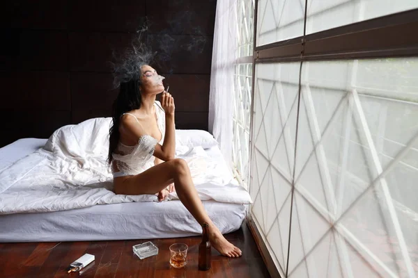 Вид збоку п'яна азіатська жінка в білій білизні, п'є і курить, тримаючи пляшку алкоголю і сидячи на ліжку в спальні — стокове фото