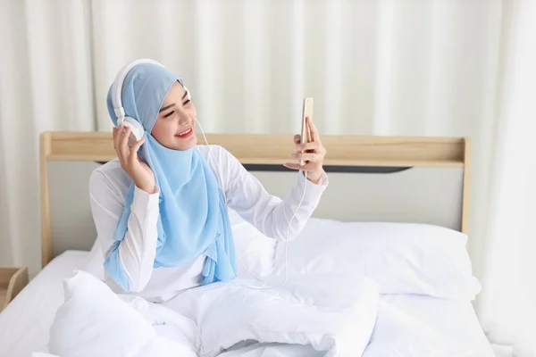 美丽的亚洲穆斯林女性穿着睡衣 在手机上观看在线故事 躺在床上 与无线网络相连 年轻可爱的戴头巾的女孩听智能手机里的音乐 — 图库照片