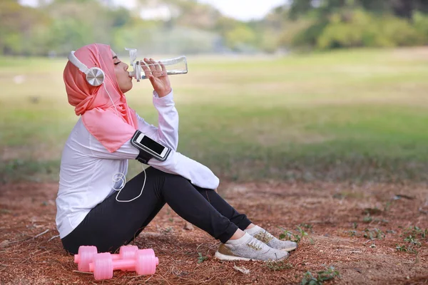 運動後に水を飲む美しいフィットネス選手アジアのイスラム教徒の女性 若いかわいい女の子はダンベルとスポーツウェアに座って ヘッドフォンは屋外でのワークアウトの後に休息を取ります 健康的でスポーツ的なコンセプト — ストック写真