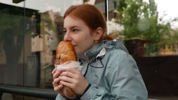 Lycklig Rödhårig Flicka Njuta Att Äta Baguette Vid Busshållplatsen — Stockvideo