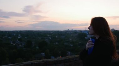 Kırmızı saçlı kız evin çatısında şehir manzaralı bir termofincan çay içiyor