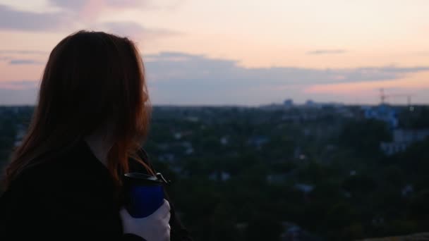 街の景色を望む家の屋根の上の赤毛の女の子は サーモカップからお茶を飲んでいます — ストック動画