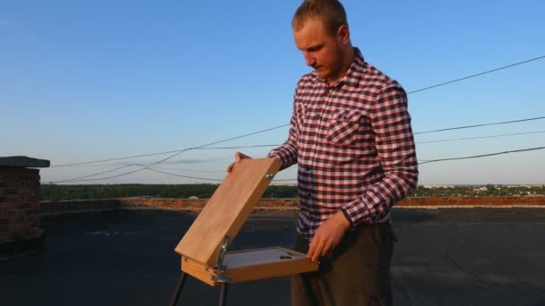 男は絵を描く 日没時の家の屋根の上で — ストック動画