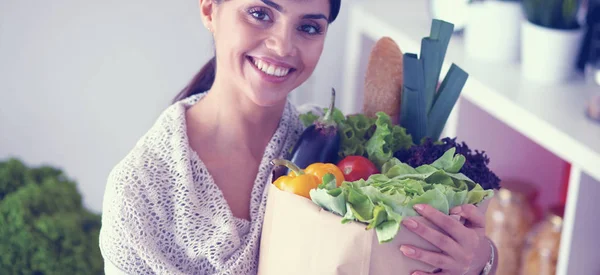 Mujer joven sosteniendo bolsa de la compra de comestibles con verduras de pie en la cocina — Foto de Stock