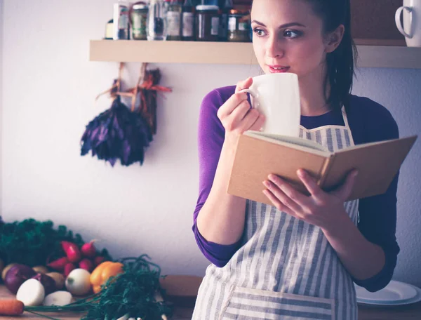 En ung kvinna som står i köket och dricker te och håller i en kokbok. Stockbild