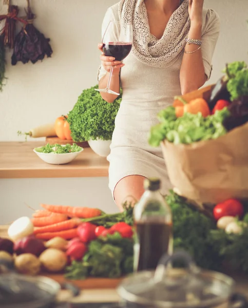 Ung kvinna skär grönsaker i köket, håller ett glas vin Stockfoto
