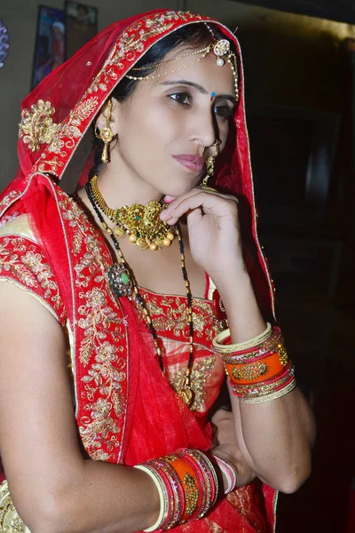 Stunning Indian bride dressed in Hindu red traditional wedding clothes sari embroidered with gold jewelry and a veil smiles with black background, looking at other side.