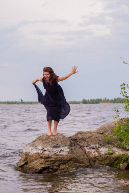 Uzun siyah elbiseli Genç çekici kız bir kayaya nehrin yanında duruyor.