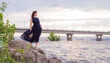 Uzun siyah elbiseli Genç çekici kız bir kayaya nehrin yanında duruyor.