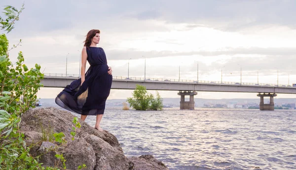 Uzun siyah elbiseli Genç çekici kız bir kayaya nehrin yanında duruyor.