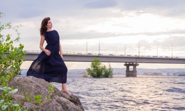 Uzun siyah elbiseli Genç çekici kız bir kayaya nehrin yanında duruyor.