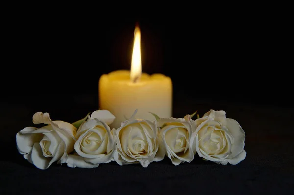 Beautiful White roses with a burning candle on the dark background. Funeral flower and candle on table against black background with copy space. Funeral symbol. Mood and Condolence card concept.