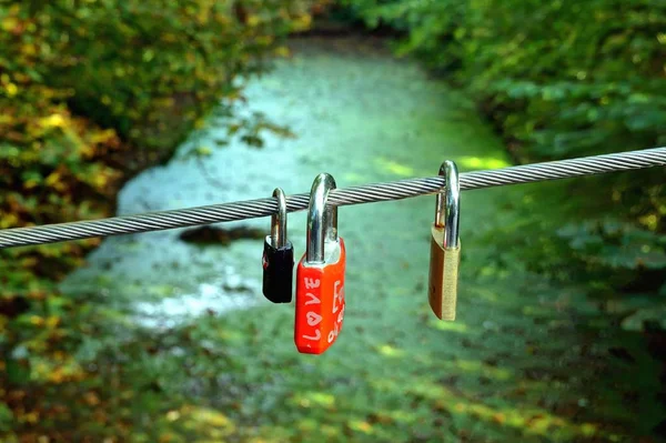 Locks of love in the shape of a heart hang on a steel cable of a pedestrian bridge, green water, river background. Padlocks, love locks lettered with love