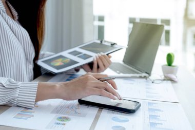 İş kadını el bilgisayarı ile akıllı telefon kullanır ve ofisteki dizüstü bilgisayarla iş hedeflerini belirlemek ve yeni hedefe ulaşmayı planlamak için dosyayı analiz eder..
