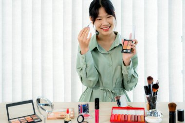 Güzel Asyalı kadın vlogger video kaydederken kozmetik makyaj ürünleri gösteriyor ve dijital akıllı telefonuyla güzellik blogu için tavsiye ediliyor.