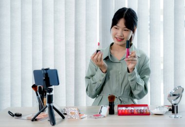 Güzel Asyalı kadın vlogger video kaydederken kozmetik makyaj ürünleri gösteriyor ve dijital akıllı telefonuyla güzellik blogu için tavsiye ediliyor.