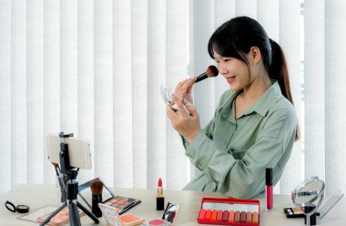 Güzel Asyalı kadın vlogger video kaydederken kozmetik makyaj ürünleri gösteriyor ve dijital akıllı telefonuyla güzellik blogu için tavsiye ediliyor.