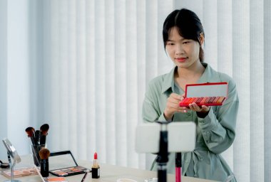 Güzel Asyalı kadın vlogger video kaydederken kozmetik makyaj ürünleri gösteriyor ve dijital akıllı telefonuyla güzellik blogu için tavsiye ediliyor.