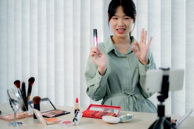 Güzel Asyalı kadın vlogger video kaydederken kozmetik makyaj ürünleri gösteriyor ve dijital akıllı telefonuyla güzellik blogu için tavsiye ediliyor.