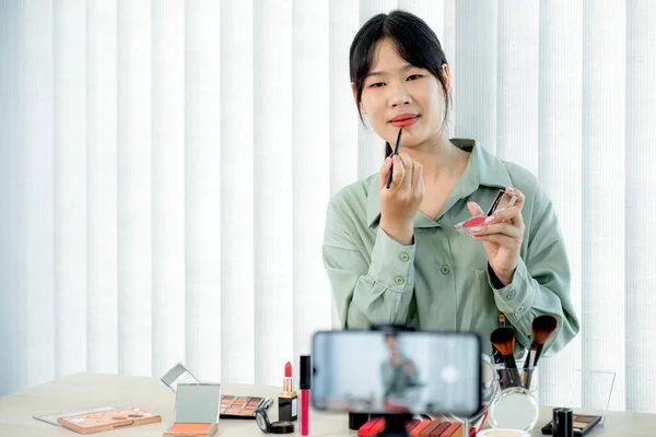 Beautiful Asia female vlogger is showing cosmetics makeup products while recording video and giving recommended for her beauty blog with digital smartphone.