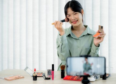 Güzel Asyalı kadın vlogger video kaydederken kozmetik makyaj ürünleri gösteriyor ve dijital akıllı telefonuyla güzellik blogu için tavsiye ediliyor.