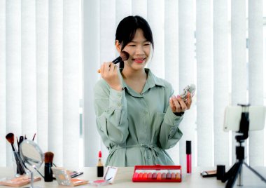 Güzel Asyalı kadın vlogger video kaydederken kozmetik makyaj ürünleri gösteriyor ve dijital akıllı telefonuyla güzellik blogu için tavsiye ediliyor.