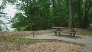 Gölün kenarındaki parkta ızgaralar ve çimento piknik masaları. Ormanın kırsal kesiminde, bulutlu bir sonbaharın ilk günlerinde her piknik alanına kaldırım döşenmiş.