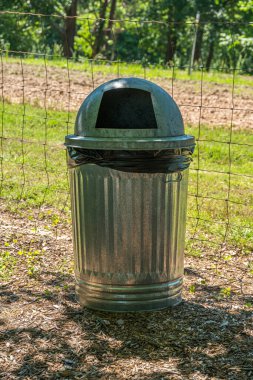 Parlak, yeni bir çöp tenekesi. Kubbe kapağı ve plastikten siyah bir çanta. Güneşli bir günde, yaz mevsiminde bir parkta.
