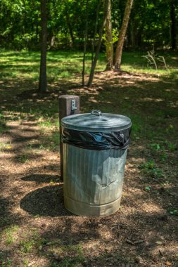Yazın güneşli bir günde, gölgeli ormanlık alanda, parkın birinde, üzerinde plastik kapaklı metal bir çöp tenekesi.