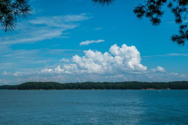 Yazın güneşli bir günde, arka planda ormanlık arazinin kıyısında birkaç tekneyle gölün üzerinde büyük, tüylü, beyaz bir bulut.